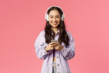 具有吸引力的现代和时尚棕褐色韩国人肖像使用手机的耳中女学生监听音乐为出去制作新的播放列表站着粉红背景具有吸引力的现代和时尚棕色韩图片