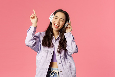 有吸引力的无照料亚洲女孩冷却和振动的肖像在耳机中听音乐在努力唱并达到歌的最高音调时举起手指粉红色背景有吸引力的无照料亚洲女孩冷却图片