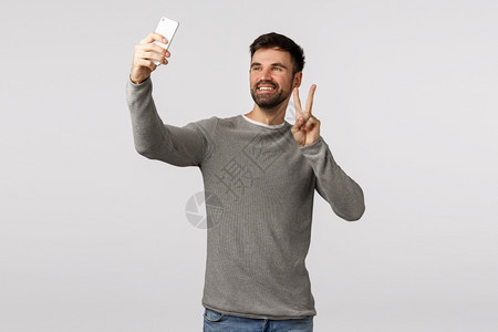 乐观而谨慎的快乐有爱心带着灰色毛衣胡子的有吸引力男用智能手机向上拍自的用智能手机向上拍的用和平信号笑的用朋友视频电话在网上打的用图片
