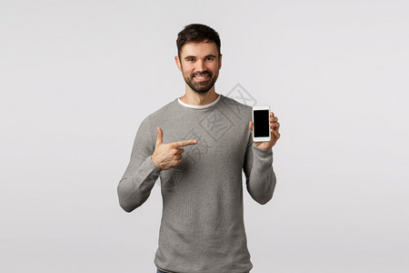 穿着灰色毛衣的英俊自信和可爱的男穿着灰色毛衣的胡子促进应用程序过滤装置指着智能手机显示笑起来做出正确的选择白背景穿着灰色毛衣宣传图片