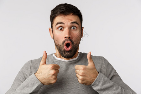 令人惊讶和着迷的帅的男穿着灰色毛衣举起拇指用惊人和吓的气喘折叠