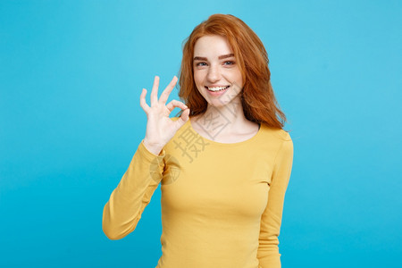 迷人的姜红发女孩头部肖像雀斑笑着用手指做一个好标记蓝色的面部背景复制空间图片