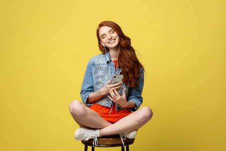 生活方式概念长卷发红的漂亮女孩在工作室的手机上听音乐坐在生动黄色背景的木椅上图片