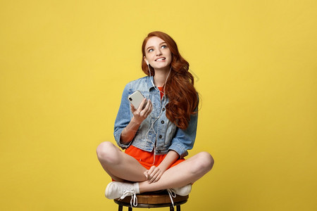 生活方式概念长卷发红的漂亮女孩在工作室的手机上听音乐坐在生动黄色背景的木椅上图片