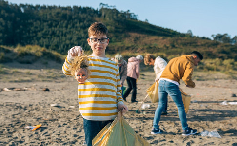 男孩用令人作呕的脸露洋娃头而志愿者团体清理海滩男孩在清理海滩时展示洋娃头图片