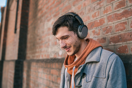 年轻拉丁男子的肖像听音乐用耳机对着砖墙城市概念图片