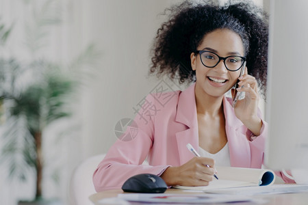 拥有卷发型的幸福非洲女写一些信息通过手机说话在办公室背景模糊的情况下坐在办公桌前图片