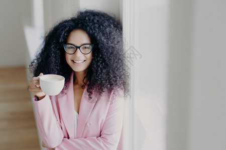 在室内拍一张照片快乐的卷头发美籍女士有咖啡休息喝着白酒穿光观眼镜正规服装站在白墙附近商业领域工作图片
