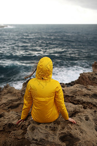 身着黄色雨衣坐在悬崖上看着海浪的大同时也享受着美丽的海景在雨天中下云雾的春季天气中岩石海滩上图片