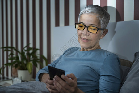 睡前用智能手机把眼镜放在床上晚用智能手机把眼镜用蓝色灯挡住的老年妇女背景图片