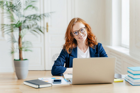 成功商业女人的红发照片戴光学眼镜进行视力矫正坐在台式桌面上坐在打开的笔记本电脑前思考组织计划周围都是纸文件图片