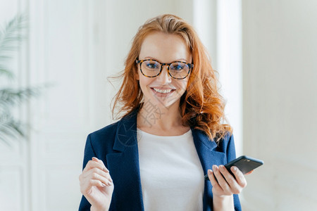 照片快乐的女领袖照片有姜头发戴眼镜更新现代手机应用程序穿优雅的衣服在现代办公室装扮阅读商业伙伴发来的信息图片