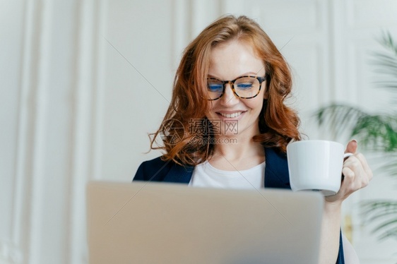穿着光眼镜喝热咖啡或茶专注在笔记本电脑线采购愉快地微笑远距离工作自由职业者书票图片