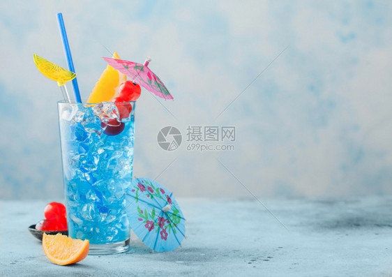 高球玻璃中的蓝色环礁湖夏季鸡尾酒配有甜樱桃和橙色切片蓝桌底有雨伞图片