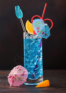 蓝色环礁湖夏季鸡尾酒在高球玻璃甜鸡尾酒樱桃和橙色切片雨伞在黑桌背景图片