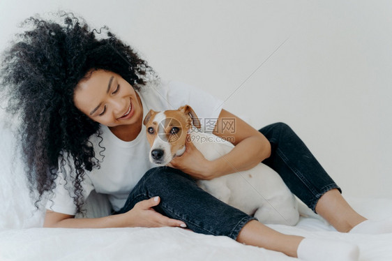 可爱的卷女人横向镜头拥抱最喜爱的狗穿whate衬衫牛仔裤和袜子一起享受时间坐在床上美好的一天开始快乐的女士在卧室和宠物玩耍图片