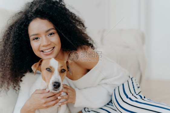 迷人的深色皮肤女形象穿着化妆品有粗糙的卷发笑容愉快拥抱的狗穿着时装享受甜蜜的时光与小狗在一起家图片