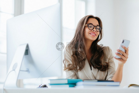 身着公司服装微笑着女士通过智能手机观看有趣的视频坐在办公室的桌面上使用计算机工作享受程序为项目浏览信息戴眼镜图片