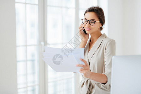 正规服装妇女打给商业伙伴的电话讨论金融启动项目阅读报纸信息戴光眼镜站在办公室国际司工作图片