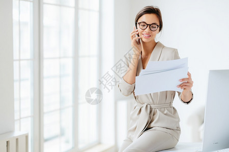 具有吸引力的女商人阅读文件或业与伙伴通过电话交谈在会前审查文书工作在会前审查文件工作坐在桌面上快乐笑与文件工作图片
