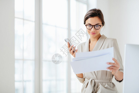 以文件为焦点的满意女雇员阅读编写的报告仔细阅读编写的报告持有现代智能手机戴透明的眼镜并穿上商业西装而不是办公室内部每月收入检查图片