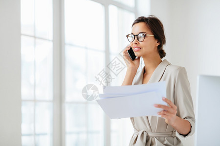 商业女人站在镜头对面持有纸文件电话交谈编写财务报告在工作空间姿势戴光学眼镜和正式西装图片