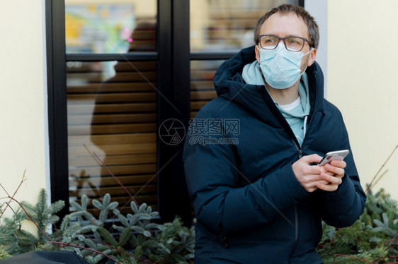 欧洲青年男子在城市街道上行走持有移动电话阅读有关冠状症的信息有在线通户外张贴保健安全概念图片