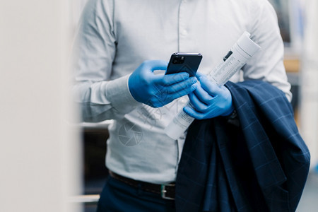 在现代智能手机上以优雅服装类型信息发布无法辨认的商人在大流行病期间佩戴保护一次手套将报纸和黑夹克卷起来在网上看新闻图片