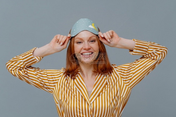 红头发正年轻女子戴眼罩睡在头上高兴地微笑穿着家庭服装晚上做甜美的梦远离灰色背景享受和平气氛图片