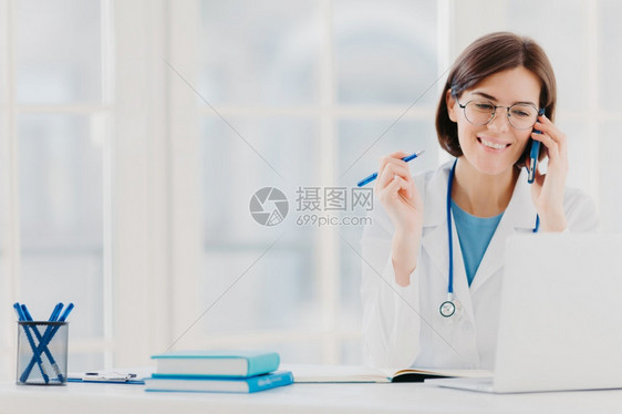 喜悦女治疗师或医生的照片与病人有电话交谈提供如何治疗疾病的建议穿白袍阅读膝上型计算机信息在私人诊所工作图片