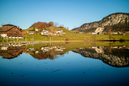 美丽的乡村景观包括传统房屋天然山丘和湖泊在清澈天空背景的蓝净水中反映出来秋天乡村风景蓝湖房屋也反映出来图片
