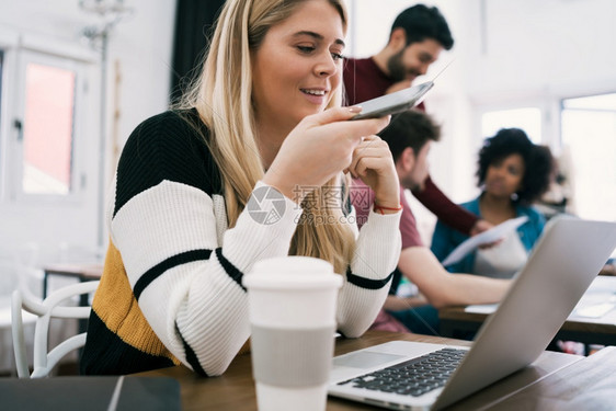 年轻商业妇女通过手机发出语音信息在工作场所使用笔记本电脑工作图片