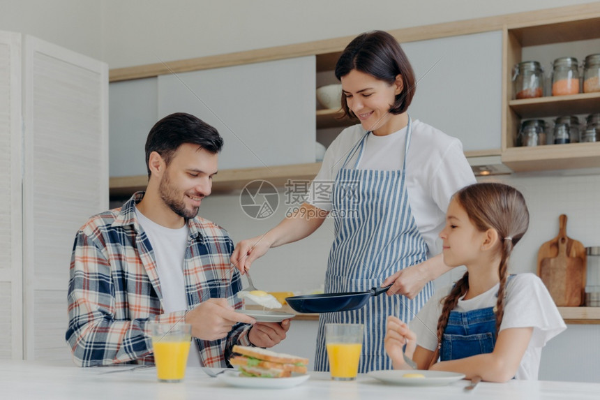 家庭主妇拿着煎锅给丈夫和女儿做饭一起吃美味的早餐坐在厨房桌边喝着果汁和三明治图片