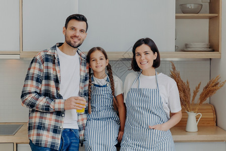 快乐的家庭有时光在一起家里的现代厨房摆姿势穿着格斗衬衫的快乐男人拿着一杯果汁小孩有猪尾辫漂亮的家庭主妇在围裙里和父母在一起的女孩图片