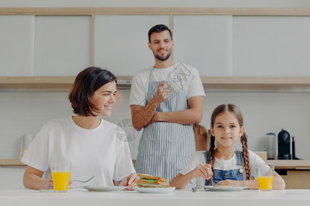 可爱的小孩和妈一起吃早餐坐在厨房桌前吃着美味的饭父亲站在背景穿围裙喝着咖啡图片