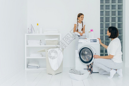 快乐的女孩坐在洗衣机上与母亲愉快地交谈剂图片