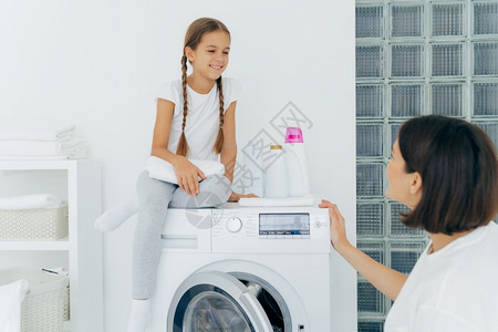 可爱的小女孩有猪尾辫在洗衣机顶上摆着姿势拿白色软毛巾高兴地看着母亲在洗完衣服后谈计划图片