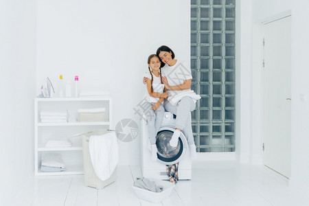 快乐的母亲和女儿有乐趣拥抱和表达彼此的爱穿着便衣坐在洗机上准备在白色宽敞的洗衣房里用脏服泡浴图片
