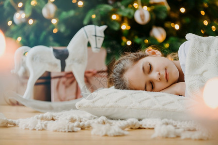 穿着白毛衣的放松女孩横向拍摄做着愉快的梦躺在装饰圣诞树下玩具摇滚乐手和礼品盒周围孩子在家舒适的假日家庭气氛图片