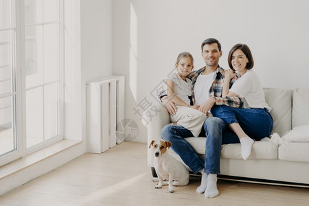幸福的男子拥抱女儿和妻坐在空的房间里坐着舒适的白色沙发他们的宠物坐在地板上做家庭肖像供人们长期记忆图片