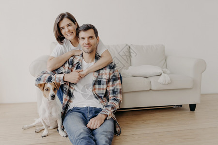 喜悦的女子相片拥抱她的丈夫在沙发上摆姿势小狗坐在地上靠近房主背景图片
