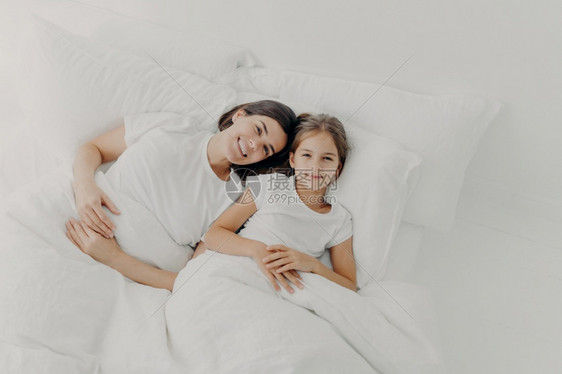 快乐的母亲和女儿在心情良好放松在床上享受舒适早快乐穿着睡衣在软枕头的白毯下睡觉时间图片