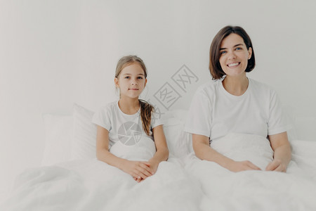 放松的妈和女儿横向镜头坐在白毯子下的床上愉快地看着镜头感到高兴花空时间在早上一起家庭时间和气氛图片