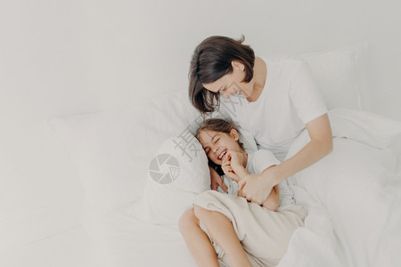 穿着睡衣的快乐有趣女孩和爱心的妈躺在床上一起玩耍享受柔软和舒适表达积极的情感图片