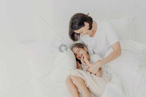 穿着睡衣的快乐有趣女孩和爱心的妈躺在床上一起玩耍享受柔软和舒适表达积极的情感图片