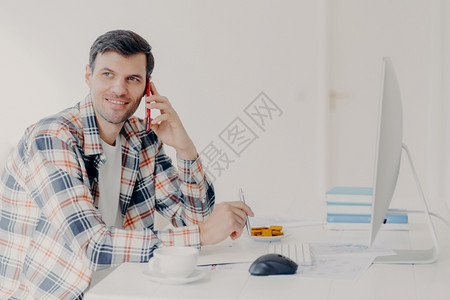 忙碌的男自由职业者横向拍摄有电话交谈穿着格衬衫写下信息与计算机坐在桌面上解决财务问题在工作场所摆姿势图片