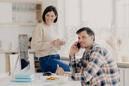 喜悦的丈夫和妻子在家管理财务审查银行账户喜悦男人把手机放在近耳边穿白衬衫喜悦女人坐在桌上喝杯子香咖啡图片