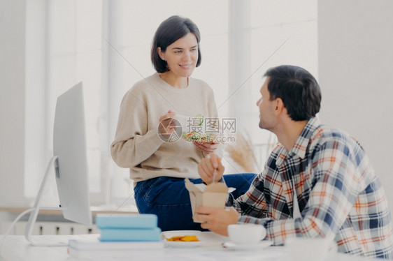 可爱的男女在吃午餐快和新鲜蔬菜沙拉在桌面上展示电脑监视器和科学文献的同时一起讨论工作问题图片
