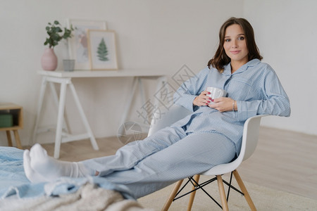 穿睡衣的放松女室内镜头坐在椅子上保持双腿在床上喝热咖啡反内脏的化妆品有吸引力的外观图片