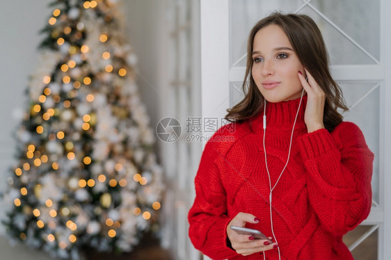 漂亮女利用手机和耳监听音乐站在家中反对圣诞节树灯背景并抄写文字空间图片